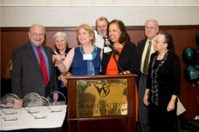 A photo of Pam Larson smiling at a podium surrounded by many Academy Members and friends.